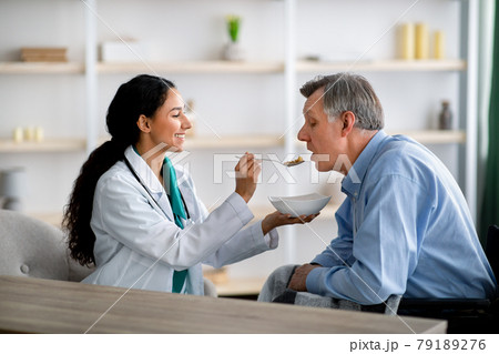 Caregiver feeding elderly man in wheelchair at home. Medical assistance for senior people with partial paralysis Caregiver feeding elderly man in wheelchair at home. Medical assistance for senior people with partial paralysis 79189276