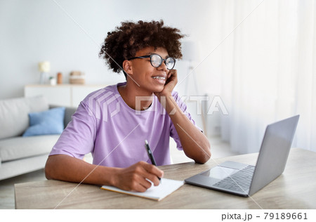 Online training. Happy black teenager studying remotely at home, writing in his notebook, looking at laptop screen Online training. Happy black teenager studying remotely at home, writing in his notebook, looking at laptop screen 79189661