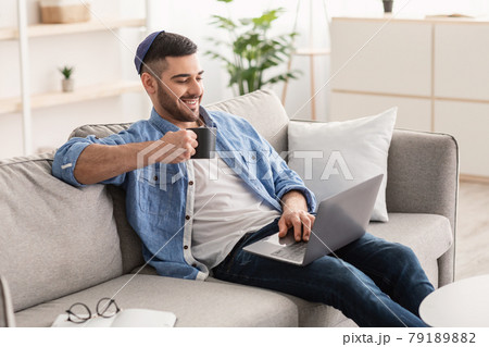 Smiling jewish man watching video on computer, drinking hot coffee Smiling jewish man watching video on computer, drinking hot coffee 79189882