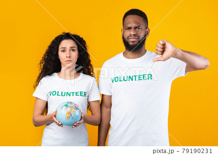 Female And Male Volunteers Holding Globe Gesturing Thumbs-Down, Yellow Background Female And Male Volunteers Holding Globe Gesturing Thumbs-Down, Yellow Background 79190231