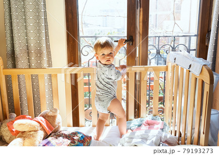 Small child opens a balcony door while standing in a crib with soft toys 79193273