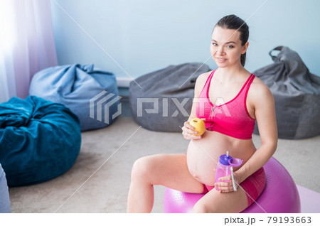 Faceless pregnant woman eating an apple after yoga. Expectant mother bites fruit and drinks water after Pilates. Girl sitting on a fitness ball. 79193663