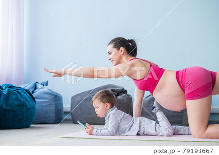 A pregnant woman in sportswear does gymnastics while her son plays in the mobile. Girl on yoga with her child. Fitness mom and baby. 79193667