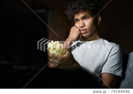 Boring film. Young guy looking bored while watching movie and holding popcorn bowl, sitting on a sofa in dark room at home Boring film. Young guy looking bored while watching movie and holding popcorn bowl, sitting on a sofa in dark room at home 79194696