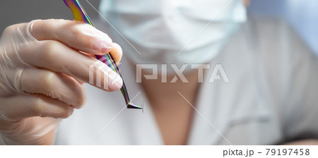 Lashmaker in a mask holds a bunch of eyelashes with tweezers. Close-up of a working tool of a master in a beauty salon 79197458