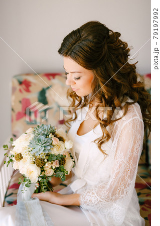 A bride in a lace robe sits on a sofa in a hotel room and holds a wedding bouquet in her hands, close-up A bride in a lace robe sits on a sofa in a hotel room and holds a wedding bouquet in her hands, close-up 79197992