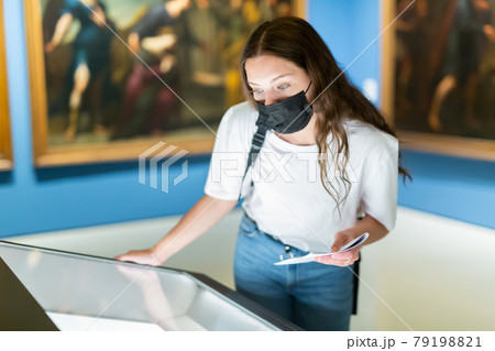 Young girl in a protective mask in the museum looks at the exposition behind a glass case Young girl in a protective mask in the museum looks at the exposition behind a glass case 79198821