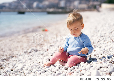Pensive kid sits on a pebble beach, looking at his feet, near the sea 79199165