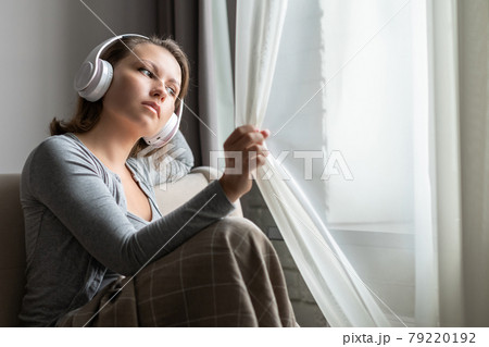 Portrait of young woman in headphones listening music sitting on sofa covered blanket and looking at window opening curtain 79220192