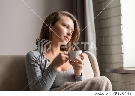 Portrait of beautiful young woman sitting on sofa covered blanket, drinking hot coffee from cup and looking at window. 79220445
