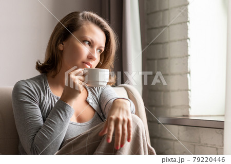 Portrait of beautiful young woman sitting on sofa covered blanket, drinking hot coffee from cup and looking at window. 79220446