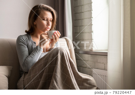 Portrait of beautiful young woman sitting on sofa covered blanket, drinking hot coffee from cup and looking at window. 79220447