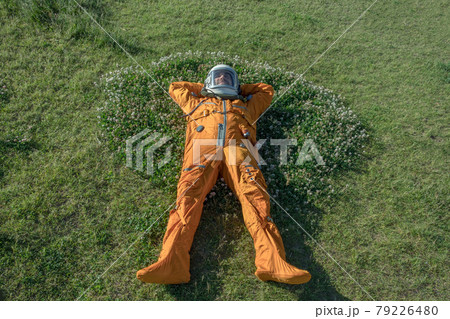 Happy cosmonaut wearing space suit and helmet having a rest while lying on green grass outdoor 79226480