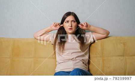 woman covers her ears and swears at the noise from neighbors while sitting on the couch 79239997