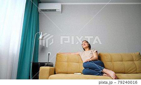 woman adjusts the air conditioner while sitting on the couch, woman adjusts the air conditioner while sitting on the couch, 79240016