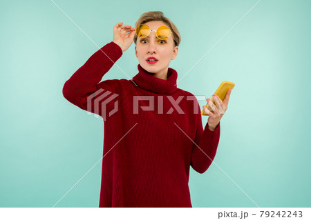 A young surprised woman in a red sweater with a smartphone on a blue background A young surprised woman in a red sweater with a smartphone on a blue background 79242243