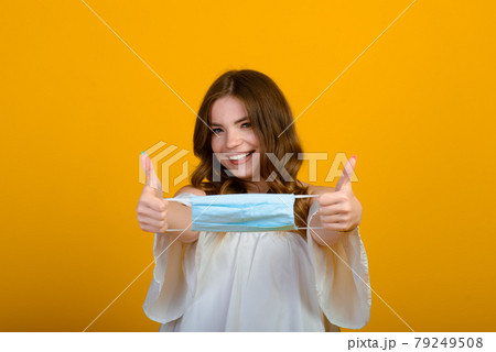 Beautiful happy young woman with face mask in hands on yellow background. COVID virus and air pollution. Recovering from illness, end of epidemic. 79249508
