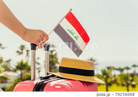 Woman with pink suitcase, hat and Iraq flag standing on passengers ladder and getting out of airplane opposite sea coastline with palm trees. 79256008