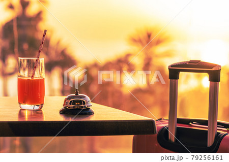 Suitcase, ringing hotel service bell with welcome drink and sea, palm tree view on sunset. Travel concept. 24-hour hotel front desk. Late check-out. 79256161