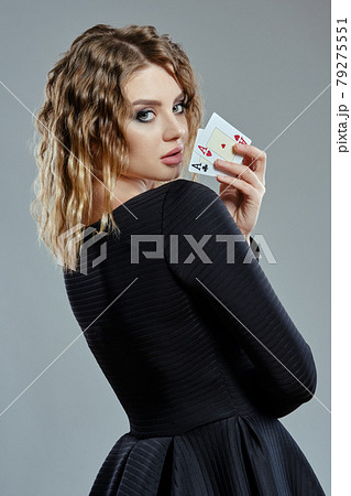 Blonde woman in black dress showing two playing cards, posing sideways against gray background. Gambling entertainment, poker, casino. Close-up. 79275551