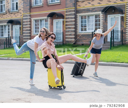 Three joyful Caucasian friends are having fun and chasing each other while riding a suitcase. Excited women go on a road trip. The brunette blonde and the redhead are going on vacation.  79282908