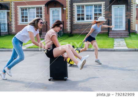 Three joyful Caucasian friends are having fun and chasing each other while riding a suitcase. Excited women go on a road trip. The brunette blonde and the redhead are going on vacation.  79282909