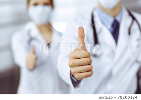 Unknown man-doctor and woman standing straight as a team and showing Ok sign with thumbs up in modern clinic. Medicine concept during Coronavirus pandemic. Covid 2019 Unknown man-doctor and woman standing straight as a team and showing Ok sign with thumbs up in modern clinic. Medicine concept during Coronavirus pandemic. Covid 2019 79300134