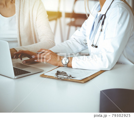 Unknown woman-doctor communicating with female patient in sunny clinic. Physician checks medical history record and exam results, close-up 79302925