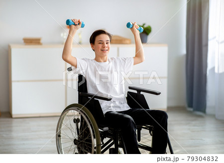 Physical rehabilitation for impaired people. Cheerful disabled teenager in wheelchair exerising with dumbbells at home 79304832