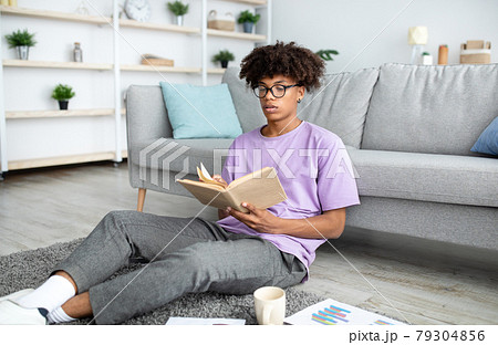 Focused black teenager reading book, sitting on floor, studying from home 79304856