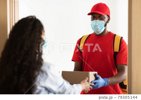 Black delivery man in mask giving box to female client 79305144