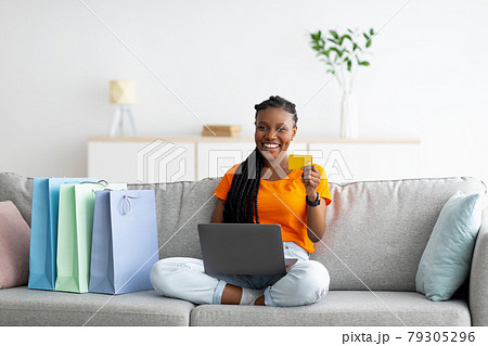 Cheerful black woman sitting on couch with laptop computer, holding credit card, making payment in online shop from home 79305296