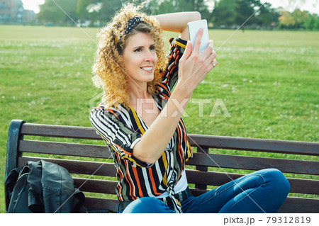 Curly hair woman making selfie photo on a mobile phone. Stylish happy smiling woman in striped multicolor shirt, posing on the background of green lawn. Summer fashion. Capture the moment. Curly hair woman making selfie photo on a mobile phone. Stylish happy smiling woman in striped multicolor shirt, posing on the background of green lawn. Summer fashion. Capture the moment. 79312819