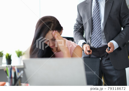 Depressed woman sits at work desk from behind man stands and unfastens belt 79318672