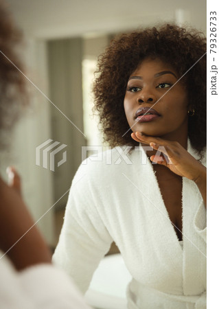 African american woman in bathroom wearing bathrobe, looking in mirror and moisturising face African american woman in bathroom wearing bathrobe, looking in mirror and moisturising face 79326733