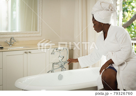 African american woman sitting in bathroom wearing bathrobe and towel, running bath 79326760