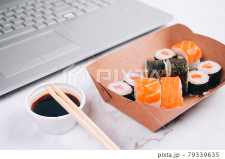 Different sushi rolls with salmon in delivery paper box on white marble table background with laptop - futomaki, uramaki and hosomaki. Online order food and delivery food concept Different sushi rolls with salmon in delivery paper box on white marble table background with laptop - futomaki, uramaki and hosomaki. Online order food and delivery food concept 79339635