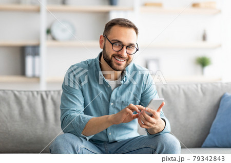 Cheerful millennial guy in glasses sitting on cozy couch with cellphone and messaging to his friend, indoors 79342843