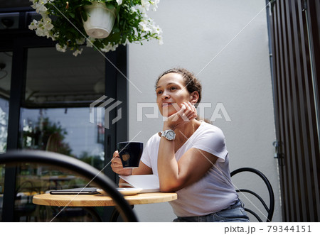 Charming woman sits in a cafe on a summer terrace and looks thoughtfully into the distance with a cup of coffee in her hand Charming woman sits in a cafe on a summer terrace and looks thoughtfully into the distance with a cup of coffee in her hand 79344151
