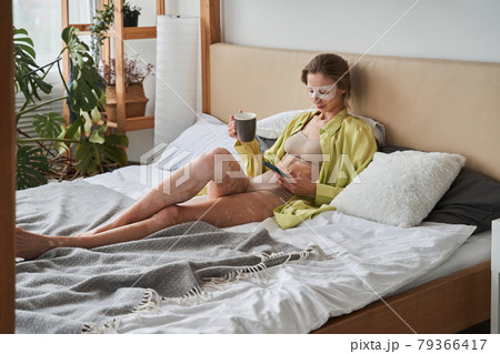 Lady in face mask drinking coffee on the bed and using her smartphone 79366417