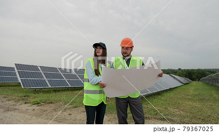 Portrait of young engineers worker is holding solar plan in hands and standing at a solar panel station. Woman and man in special uniform and protective helmet studies paper scheme. Green energy Portrait of young engineers worker is holding solar plan in hands and standing at a solar panel station. Woman and man in special uniform and protective helmet studies paper scheme. Green energy 79367025