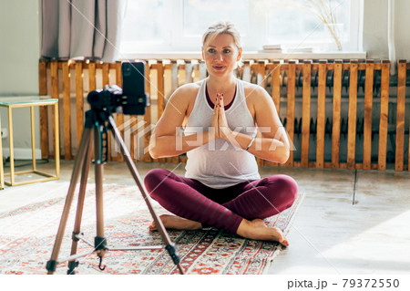 A woman sitting in a lotus position and greeting in a namaste gesture looks at the phone screen. A woman sitting in a lotus position and greeting in a namaste gesture looks at the phone screen. 79372550