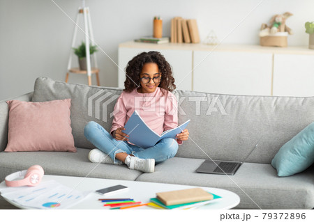 Learning and education concept. Smiling african american girl reading and doing homework, sitting on sofa with laptop Learning and education concept. Smiling african american girl reading and doing homework, sitting on sofa with laptop 79372896