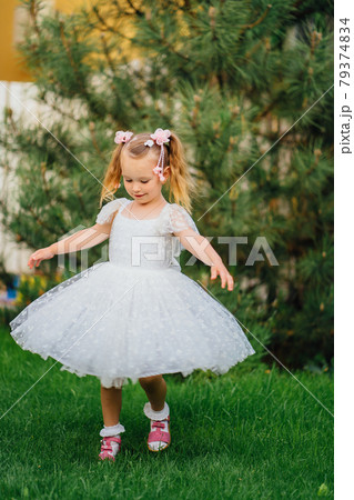 Happy little girl in white dress spinning and dancing on the lawn in the garden 79374834