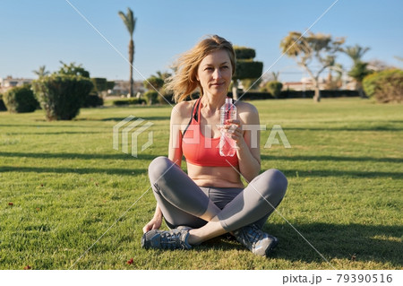 Mature woman doing morning exercises on green grass in park, with bottle of water Mature woman doing morning exercises on green grass in park, with bottle of water 79390516