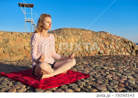 Beautiful mature woman sitting in lotus position meditating on seashore at sunset Beautiful mature woman sitting in lotus position meditating on seashore at sunset 79390543