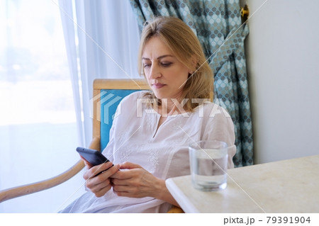 Serious middle aged woman with smartphone sitting on chair at home 79391904