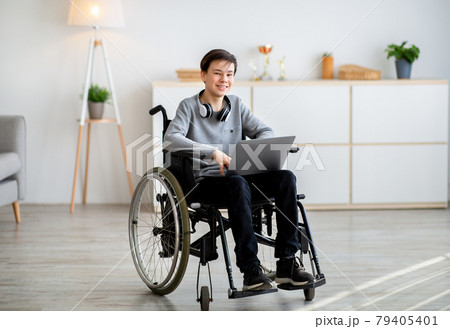 Web-based education for disabled pupils. Cheerful teen boy in wheelchair using laptop for online studies at home 79405401
