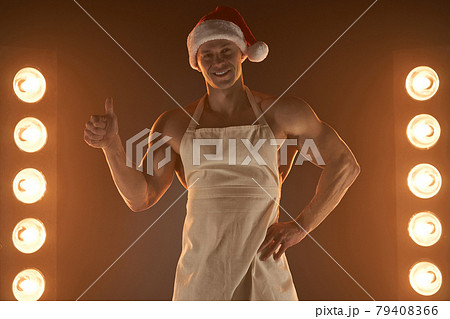 Hot Christmas dessert. Muscular man wearing apron and Santa hat showing thumb up gesture and smiling at camera, lamps illumination on background Hot Christmas dessert. Muscular man wearing apron and Santa hat showing thumb up gesture and smiling at camera, lamps illumination on background 79408366
