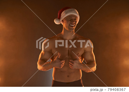 Portrait of muscular man wearing Christmas Santa hat, showing thumb up on smoky background Portrait of muscular man wearing Christmas Santa hat, showing thumb up on smoky background 79408367